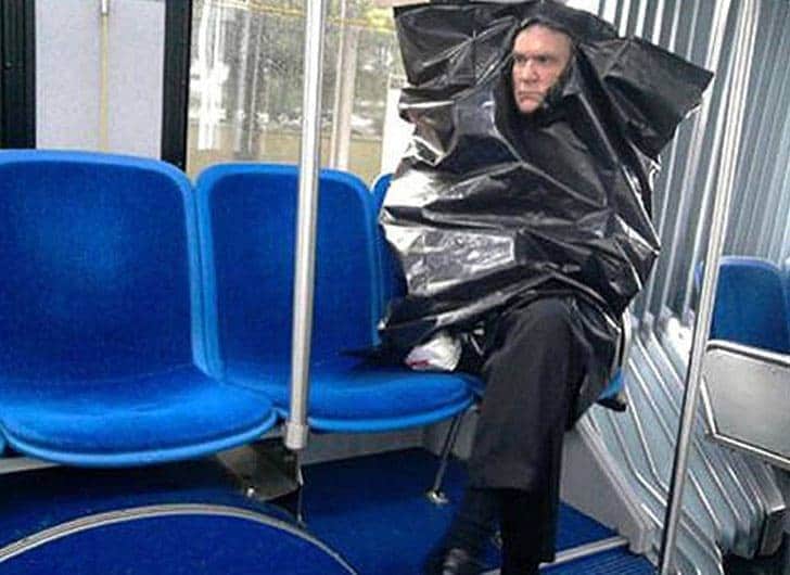 A man sits on the subway while wearing a garbage bag over his body. 